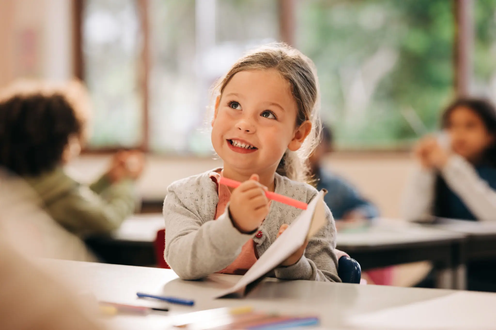 A young girl smiling while drawing in a classroom.